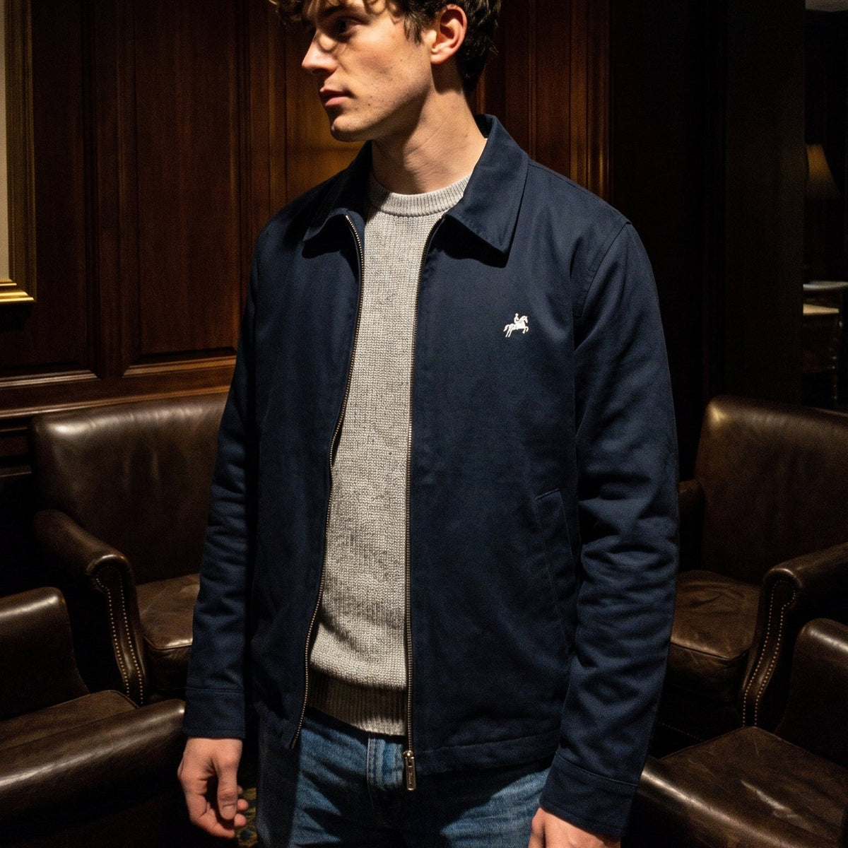 A man wearing a navy blue zip-up jacket with a small white embroidered logo on the chest, layered over a gray crewneck sweater and blue jeans, standing in a dimly lit room with dark wood paneling and leather armchairs.