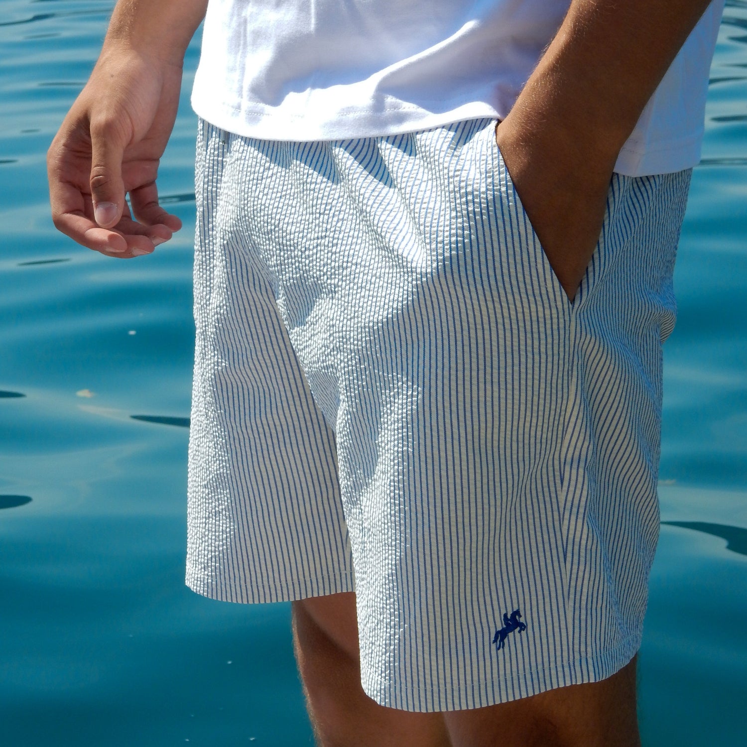 A person wearing light blue and white vertically striped textured swim shorts with a small navy logo, standing on a dock with blue water in the background.