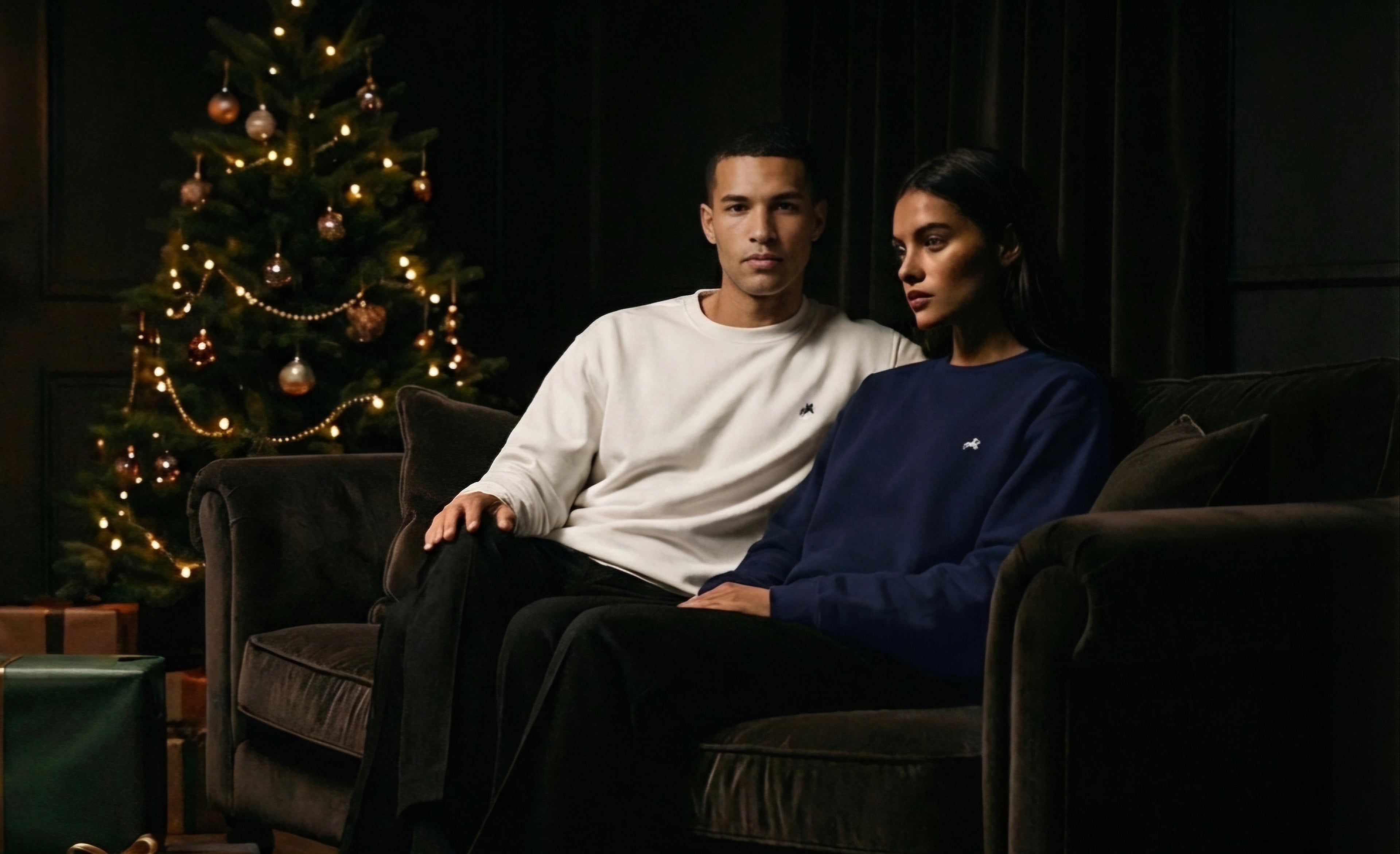 Two people sitting on a couch in a dimly lit room with a Christmas tree and presents.