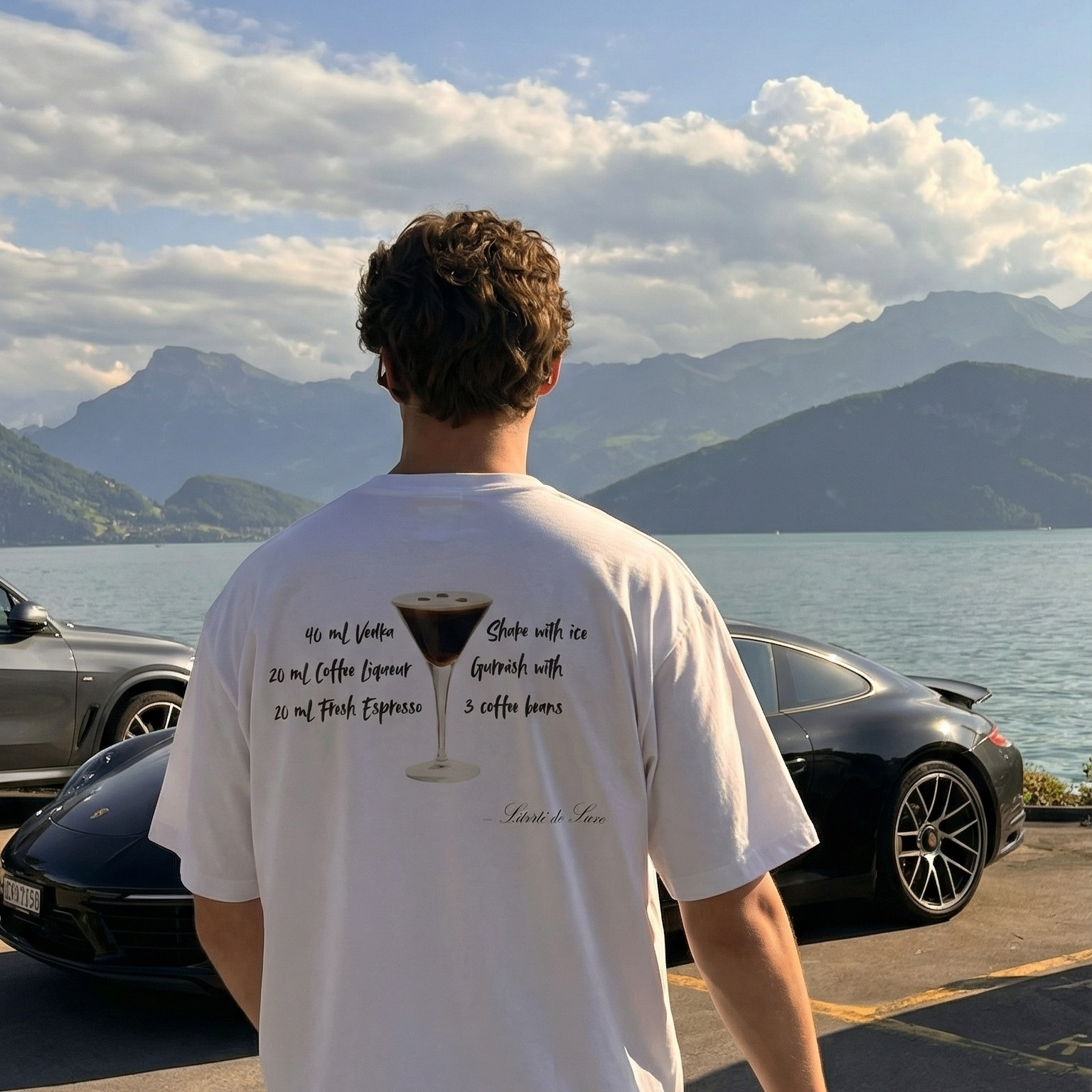 Person wearing a white t-shirt with text and graphics, standing in front of a scenic view with mountains and water.