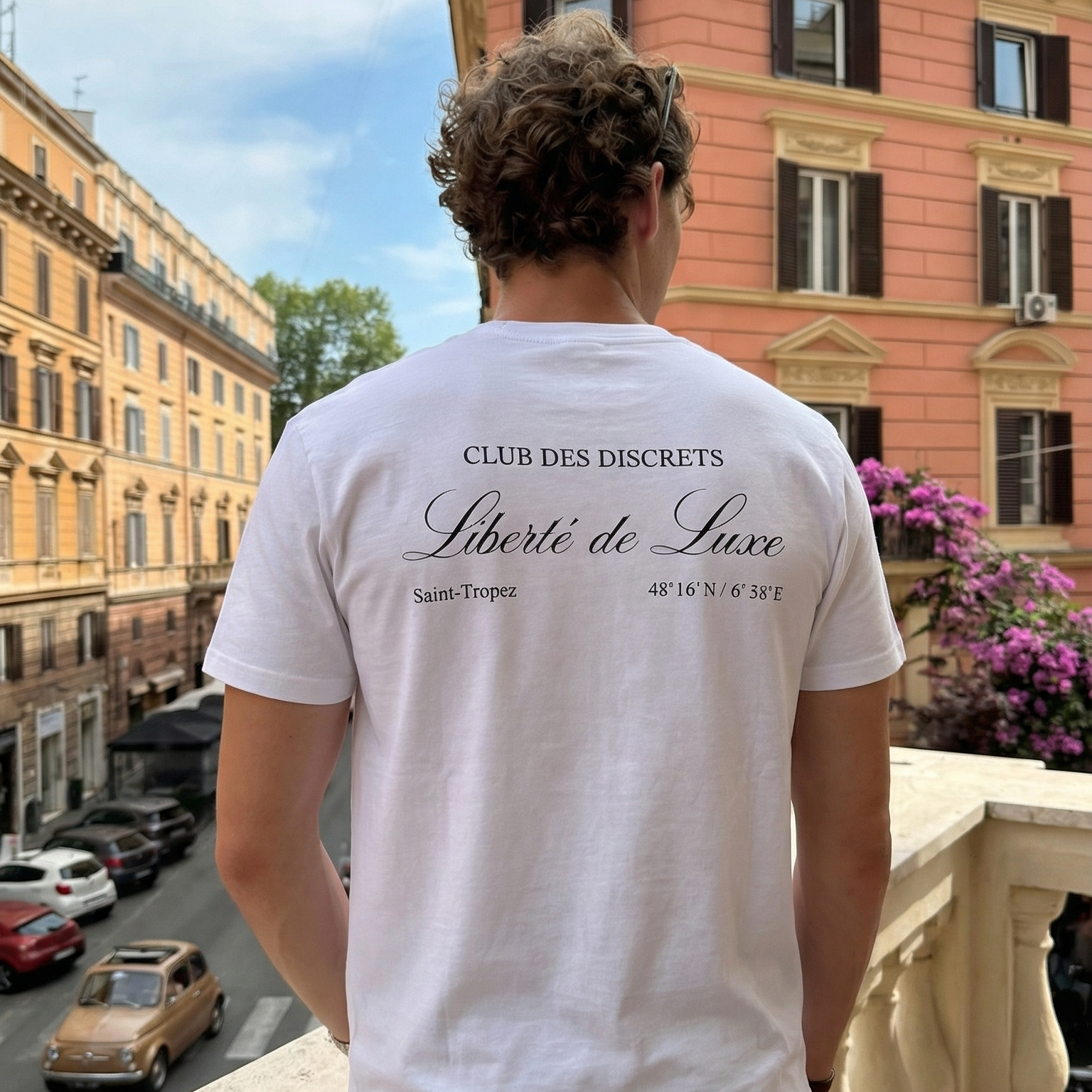 Person wearing a white t-shirt with text standing on a balcony overlooking a city street.