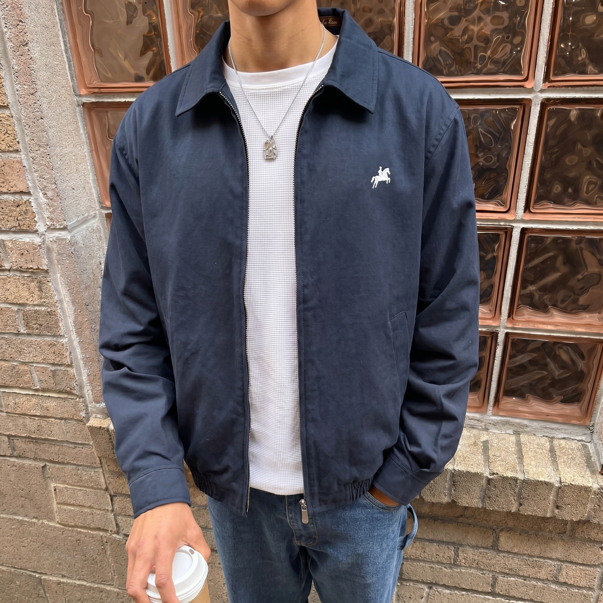 Person wearing a navy blue jacket with a logo, holding a coffee cup, standing against a brick wall.