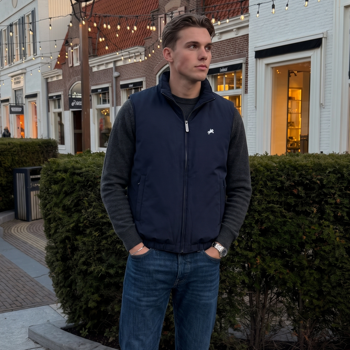 A man wearing a navy blue zip-up vest over a dark gray long-sleeve shirt, blue jeans, and white sneakers, standing outdoors on a paved street in front of European-style buildings and hedges.