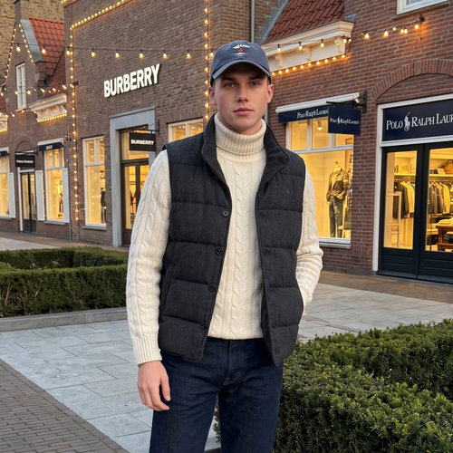 A man stands outdoors on a shopping plaza sidewalk in front of brick storefronts, wearing a cream turtleneck sweater, black quilted puffer vest, dark blue jeans, white sneakers, and a dark baseball cap.