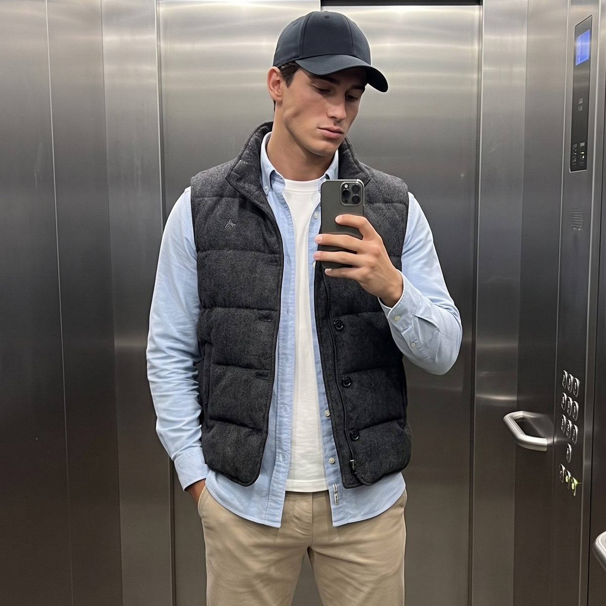 A man takes a mirror selfie in an elevator wearing a black quilted puffer vest over a light blue button-down shirt, khaki pants, and white sneakers.